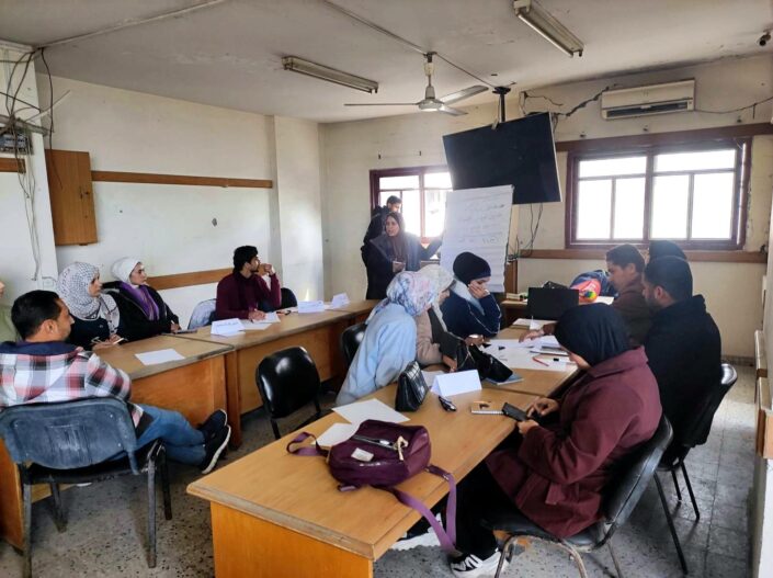 مدرّبة بجانب لوحة ورقية فيما يعمل المشاركون على أوراق حول الطاولات